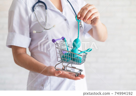 A faceless gynecologist puts a set of kegel balls into a miniature shopping cart. The doctor holds a mini trolley and device for vaginal muscles. Female health concept. 74994192
