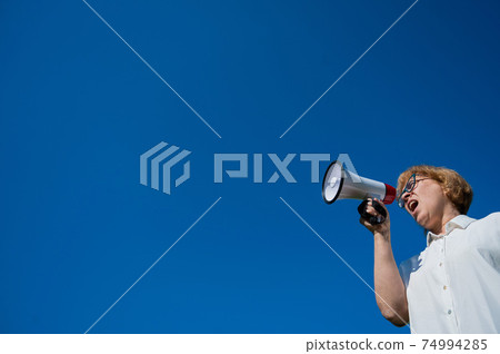 Angry senior woman in glasses shouting into a megaphone on the outside. Annoyed pensioner yells into a device amplifying sound against a blue sky. Loudspeaker in female hands. Angry senior woman in glasses shouting into a megaphone on the outside. Annoyed pensioner yells into a device amplifying sound against a blue sky. Loudspeaker in female hands. 74994285