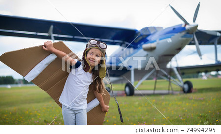 A little girl in a pilot's costume with cardboard wings runs on the lawn against the backdrop of the plane. A child in a hat and glasses dreams of flying on an airplane. A little girl in a pilot's costume with cardboard wings runs on the lawn against the backdrop of the plane. A child in a hat and glasses dreams of flying on an airplane. 74994295