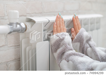 A woman wearing in pajamas warms cold hands on the heating radiator 74994299