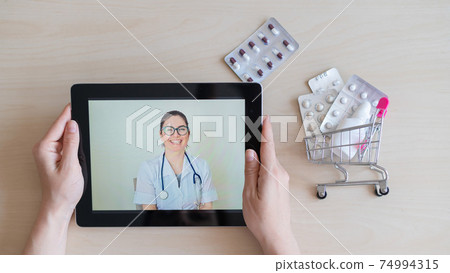 Smiling female doctor on the screen of a digital tablet. Online appointment of a general practitioner. Mini medicine trolley. Remote consultation. Smiling female doctor on the screen of a digital tablet. Online appointment of a general practitioner. Mini medicine trolley. Remote consultation. 74994315