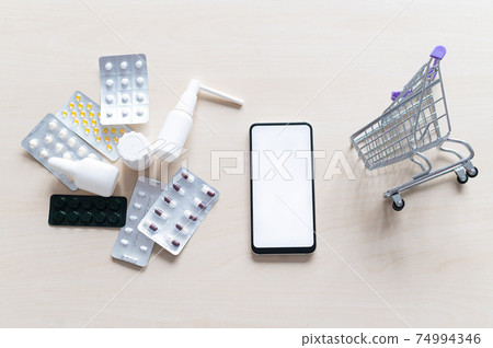 Online pharmacy concept with home delivery. Buying medicine online by phone. Mini trolley with different tablets, sprays, capsules and a smartphone with a white blank screen. Arrow made of pills. 74994346