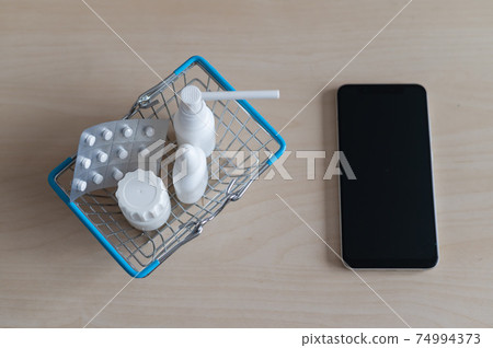 Online pharmacy. Blister with pills throat spray jar with capsules in a mini shopping basket next to a mobile phone with a black screen. Smartphone app for buying medicine. Online pharmacy. Blister with pills throat spray jar with capsules in a mini shopping basket next to a mobile phone with a black screen. Smartphone app for buying medicine. 74994373