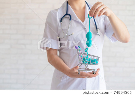 A faceless gynecologist puts a set of kegel balls into a miniature shopping cart. The doctor holds a mini trolley and device for vaginal muscles. Female health concept. 74994565