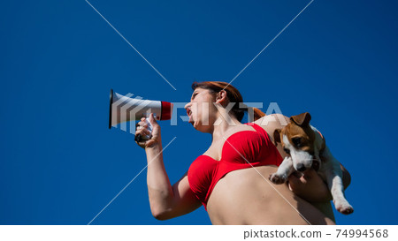 Caucasian woman in red bikini works as a lifeguard on the beach and shouts through a megaphone against a blue sky. Girl holds puppy jack russell terrier and loudspeaker. Summer vacation. Caucasian woman in red bikini works as a lifeguard on the beach and shouts through a megaphone against a blue sky. Girl holds puppy jack russell terrier and loudspeaker. Summer vacation. 74994568