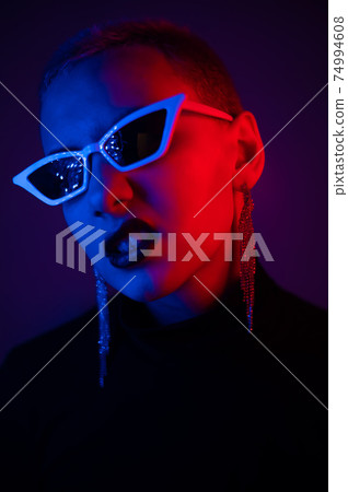 Portrait of a young woman with a short male haircut in sunglasses in neon light in the studio 74994608