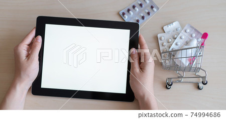 Digital tablet with a white screen in female hands and a mini cart with blisters of pills. The concept of a mobile application for the remote purchase of medicines. Online pharmacy. 74994686