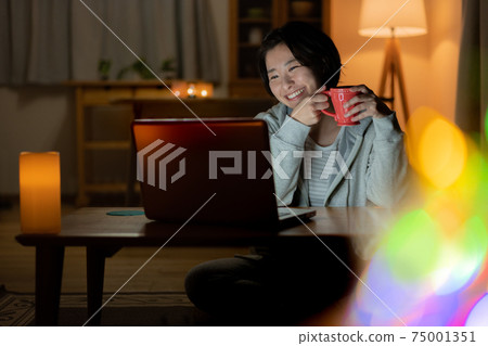 晚上看著客廳裡的一台電腦時喝一杯微笑的年輕女子 晚上看著客廳裡的一台電腦時喝一杯微笑的年輕女子 75001351