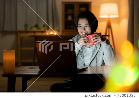 晚上看著客廳裡的一台電腦時喝一杯微笑的年輕女子 晚上看著客廳裡的一台電腦時喝一杯微笑的年輕女子 75001353