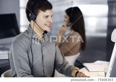 Young dark-haired guy in a green shirt and headsets is talking to a client, while sitting at the desk, working together with a female colleague in a modern office. Call center operators at work Young dark-haired guy in a green shirt and headsets is talking to a client, while sitting at the desk, working together with a female colleague in a modern office. Call center operators at work 75006363