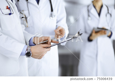 Unknown doctors, two men and a woman, discuss medical exam resoults, while standing at hospital office. Physicians using clipboards for filling up medication history records. Perfect medical service 75006439