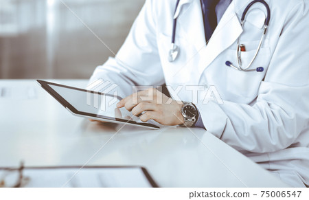 Unknown male doctor sitting and working with tablet computer in clinic at his working place, close-up. Young physician at work. Perfect medical service, medicine concept Unknown male doctor sitting and working with tablet computer in clinic at his working place, close-up. Young physician at work. Perfect medical service, medicine concept 75006547