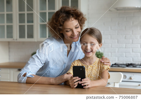Interested mom sit by table with little daughter using cellphone 75006807