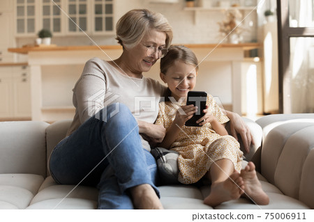 Happy girl and grandmother embrace on couch look at cell 75006911