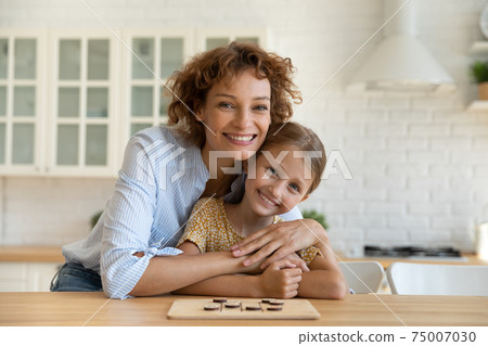 Affectionate young woman parent cuddle girl child by kitchen table Affectionate young woman parent cuddle girl child by kitchen table 75007030