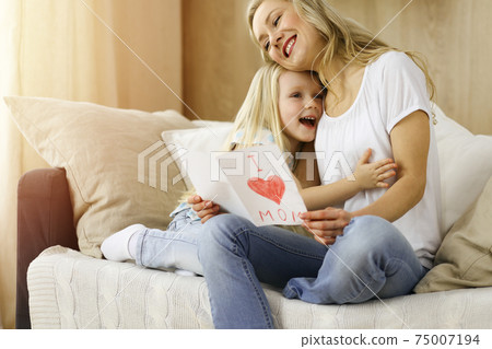Happy mother day in sunny flat. Child daughter congratulates mom and gives her postcard with heart drawing. Family concept 75007194