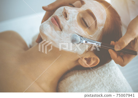 Beautiful brunette woman enjoying applying cosmetic mask with closed eyes. Relaxing treatment in medicine and spa concepts 75007941