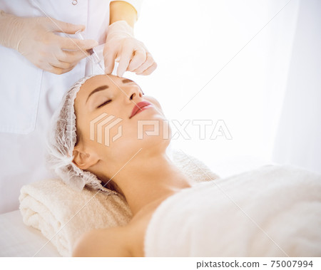 Beautiful woman receiving beauty injections with closed eyes in sunny medical center. Beautician doctor hands doing beauty procedure to female face with syringe. Cosmetic medicine and surgery concept Beautiful woman receiving beauty injections with closed eyes in sunny medical center. Beautician doctor hands doing beauty procedure to female face with syringe. Cosmetic medicine and surgery concept 75007994