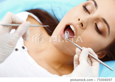 Young female patient visiting dentist office.Beautiful woman with healthy straight white teeth sitting at dental chair with open mouth during oral checkup while doctor working at teeth Young female patient visiting dentist office.Beautiful woman with healthy straight white teeth sitting at dental chair with open mouth during oral checkup while doctor working at teeth 75008097