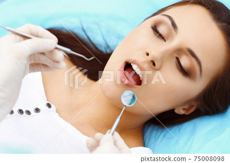 Young female patient visiting dentist office.Beautiful woman with healthy straight white teeth sitting at dental chair with open mouth during oral checkup while doctor working at teeth 75008098