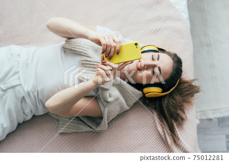 Young carefree millennial woman in headphones lying on the bed communicates on the phone and smiles. Young carefree millennial woman in headphones lying on the bed communicates on the phone and smiles. 75012281