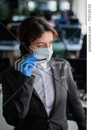 Female call center operator talking to a customer. Woman in mask and gloves at the workplace in the office. Social distance and safety measures to prevent infection. Female call center operator talking to a customer. Woman in mask and gloves at the workplace in the office. Social distance and safety measures to prevent infection. 75018539