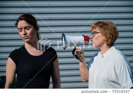 The conflict of generations. An old woman yells into a megaphone at her middle-aged daughter against gray background. Quarrel between an elderly mother and daughter. 75018547