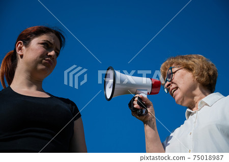 The conflict of generations. An emotional elderly woman shouting at her daughter in a megaphone. An elderly mother swears at a middle-aged woman on a loudspeaker on a blue background. 75018987
