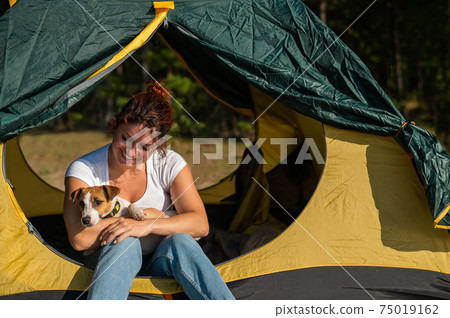 Beautiful red-haired woman is resting in nature with her pet. A girl sits in a tourist tent and hugs a Jack Russell Terrier in the forest. The dog and its female owner at the campsite. Beautiful red-haired woman is resting in nature with her pet. A girl sits in a tourist tent and hugs a Jack Russell Terrier in the forest. The dog and its female owner at the campsite. 75019162
