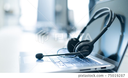 Call center operator desktop. Close-up of a headset on a laptop. Help desk. Workplace of a support service employee. Headphones with a microphone for voip on a computer keyboard. 75019287