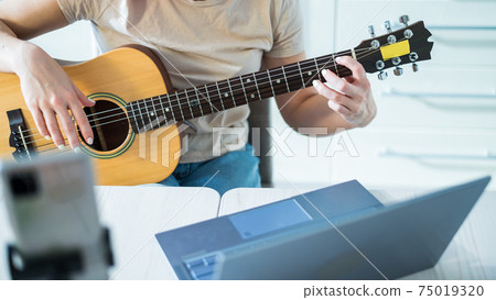 A woman sits in the kitchen during a remote acoustic guitar lesson. A girl learns to play the guitar and watches educational videos on a laptop A woman sits in the kitchen during a remote acoustic guitar lesson. A girl learns to play the guitar and watches educational videos on a laptop 75019320