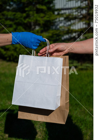 A faceless courier in gloves hands an eco friendly paper bag to a customer in a park. The concept of donation of essential products for the elderly during the coronavirus epidemic. 75019932