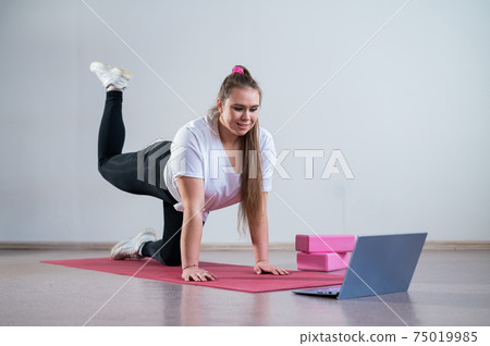 Young woman doing sports exercises at home. The girl is engaged in fitness at home on the mat and watches a training video on a laptop Young woman doing sports exercises at home. The girl is engaged in fitness at home on the mat and watches a training video on a laptop 75019985