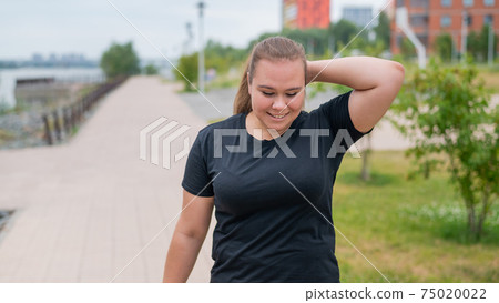 An obese young woman jogging outdoors. Fat beautiful smiling girl in a black tracksuit is engaged in fitness for weight loss on the waterfront. A woman runs on a summer day. An obese young woman jogging outdoors. Fat beautiful smiling girl in a black tracksuit is engaged in fitness for weight loss on the waterfront. A woman runs on a summer day. 75020022