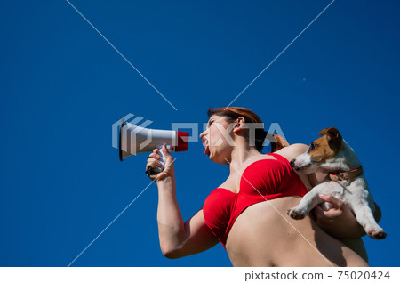 Caucasian woman in red bikini works as a lifeguard on the beach and shouts through a megaphone against a blue sky. Girl holds puppy jack russell terrier and loudspeaker. Summer vacation. 75020424
