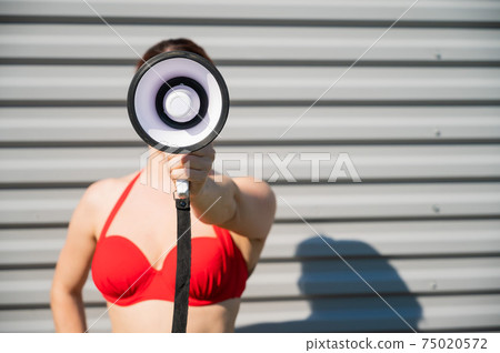 Close-up portrait of a woman in a red bikini holds a megaphone. A girl in a swimsuit makes an announcement on the speaker against the background of a steel sheet. Close-up portrait of a woman in a red bikini holds a megaphone. A girl in a swimsuit makes an announcement on the speaker against the background of a steel sheet. 75020572