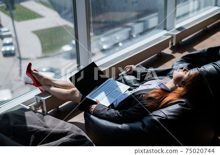 Caucasian woman fell asleep while sitting on bean bag and working on laptop in modern office 75020744