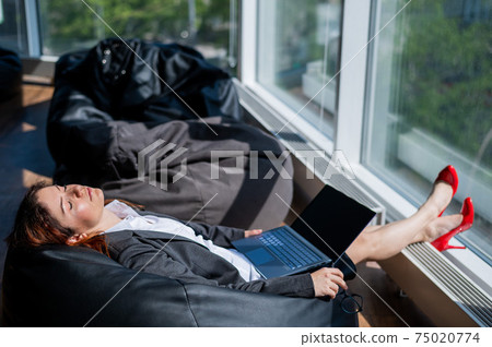 Business lady sleeps sitting in a modern office comfortable bob chair bag. A woman in a suit fell asleep at work on a laptop. Caring for the convenience and comfort of employees. 75020774