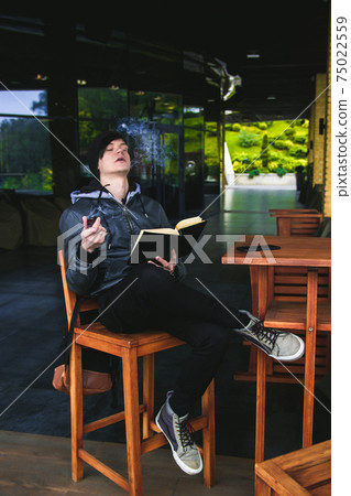 Young black haired man witn piercing reeding a book and smoking tabacco pipe in outdoor cafe. Young black haired man witn piercing reeding a book and smoking tabacco pipe in outdoor cafe. 75022559