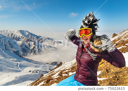 Emotional traveler young woman with windy hair smiling, sitting on top of sunny mountains. Funny woman in mohawk hat. 75022615