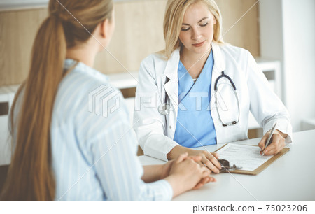 Woman-doctor and patient sitting and talking in hospital or clinic. Blonde therapist is cheerfully smiling. Medicine concept Woman-doctor and patient sitting and talking in hospital or clinic. Blonde therapist is cheerfully smiling. Medicine concept 75023206