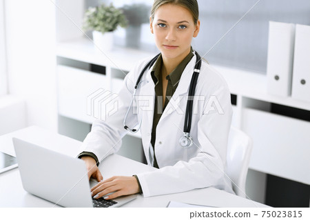 Woman-doctor typing on laptop computer while sitting at the desk in hospital office. Physician at work. Data in medicine and healthcare Woman-doctor typing on laptop computer while sitting at the desk in hospital office. Physician at work. Data in medicine and healthcare 75023815