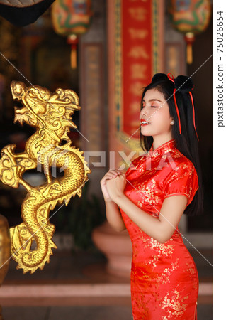 Beautiful young Asian woman in red Chinese dress traditional cheongsam qipao with gesture of congratulation and happy smiling face, she standing in front of ancient temple (Chinese new year concept) 75026654