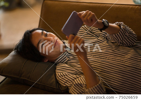 A man lying on the sofa and looking at his smartphone A man lying on the sofa and looking at his smartphone 75026659