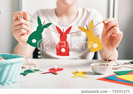 DIY Easter home decor. Happy boy make garland of colored paper, twine and pom-poms. Selective focus DIY Easter home decor. Happy boy make garland of colored paper, twine and pom-poms. Selective focus 75035375