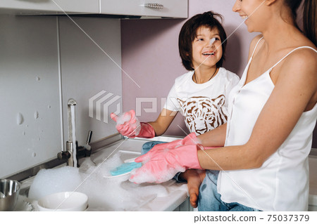 housewife mom in pink gloves washes dishes with her son by hand in the sink with detergent. A girl in white and a child with a cast cleans the house and washes dishes in homemade pink gloves.A child 75037379