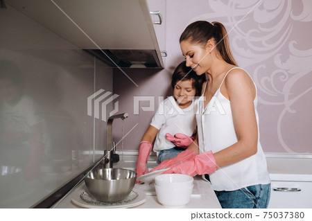 housewife mom in pink gloves washes dishes with her son by hand in the sink with detergent. A girl in white and a child with a cast cleans the house and washes dishes in homemade pink gloves.A child 75037380