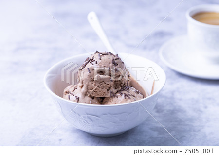 Chocolate ice cream with chocolate sprinkle in a white bowl on a light background. Three balls of homemade ice cream and a cup of coffee. Close-up. Chocolate ice cream with chocolate sprinkle in a white bowl on a light background. Three balls of homemade ice cream and a cup of coffee. Close-up. 75051001