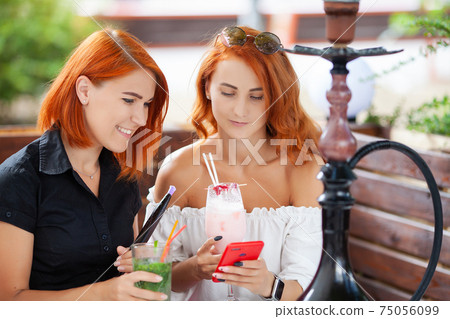 Two young women smoke a hookah and enjoy cocktails in cafe on the street Two young women smoke a hookah and enjoy cocktails in cafe on the street 75056099