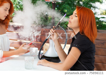 Two young women smoke a hookah and enjoy cocktails in cafe on the street Two young women smoke a hookah and enjoy cocktails in cafe on the street 75056111